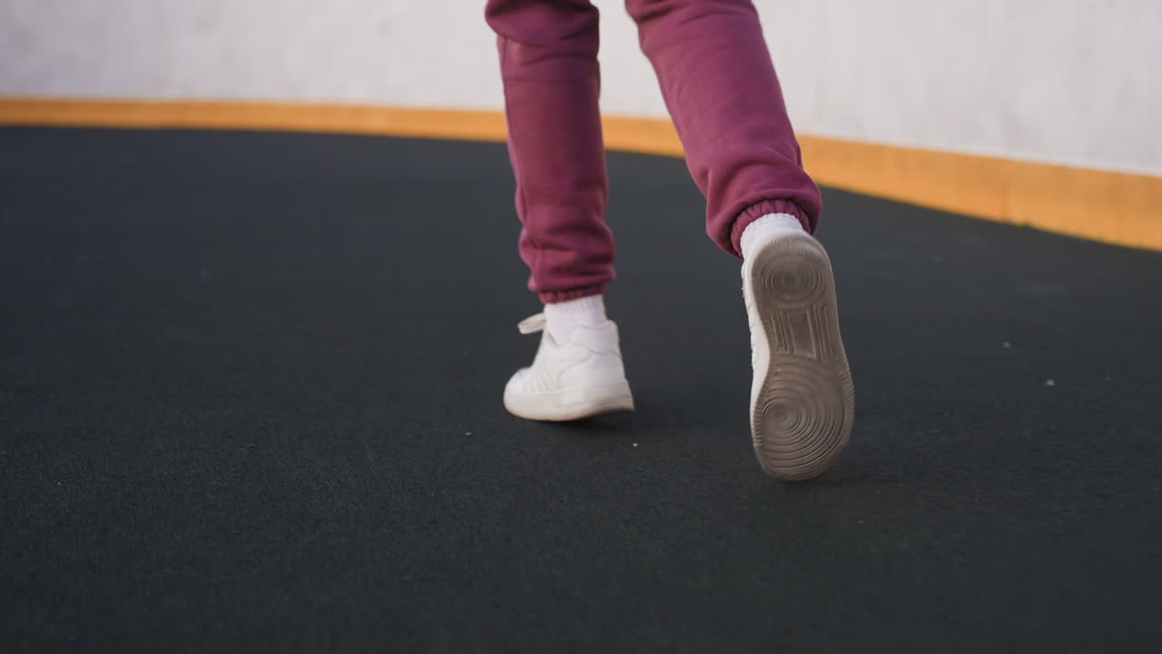 Leg view of exercise lover jogging around curved outdoor barbed wire sports court under pastel dusk sky, white sneakers stepping on smooth asphalt near yellow barrier wall with chain link fence