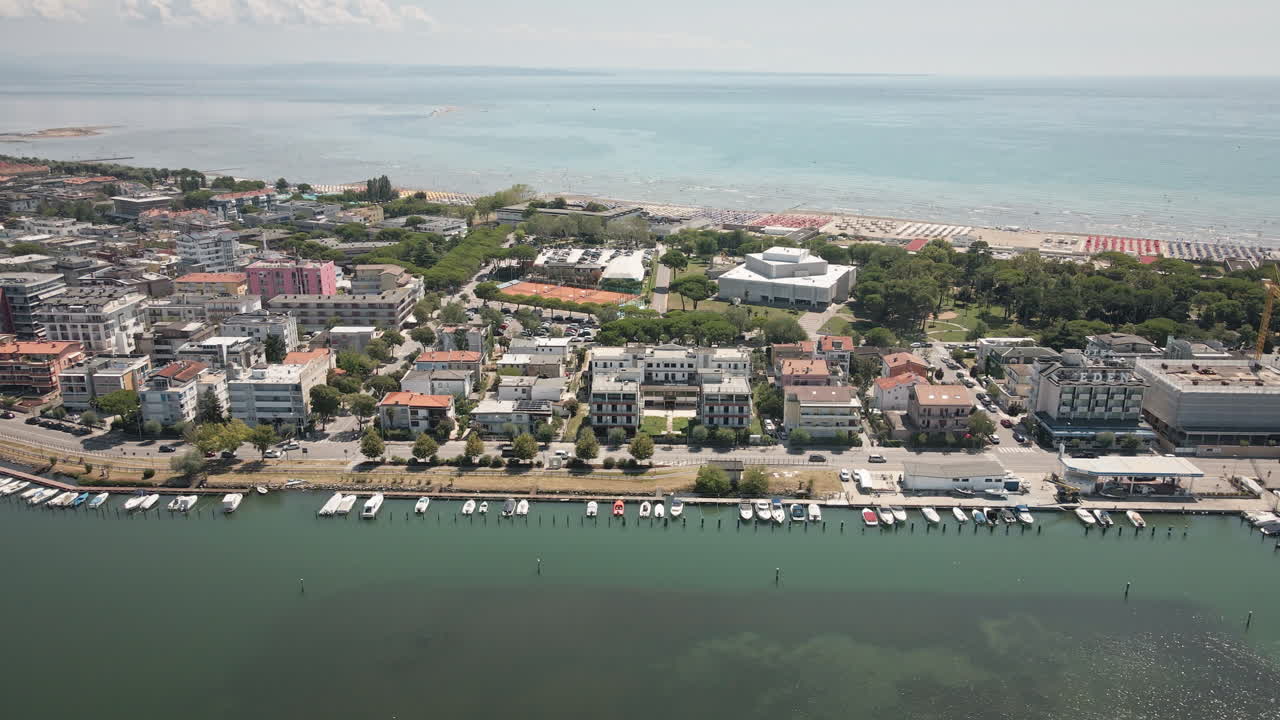 Un drone 4K UHD fluido si muove a destra lungo la città di Grado, mostrando edifici residenziali, il porto turistico con le barche e la vasta laguna — perfetto per promozioni di viaggio, b-roll, paesaggi urbani.