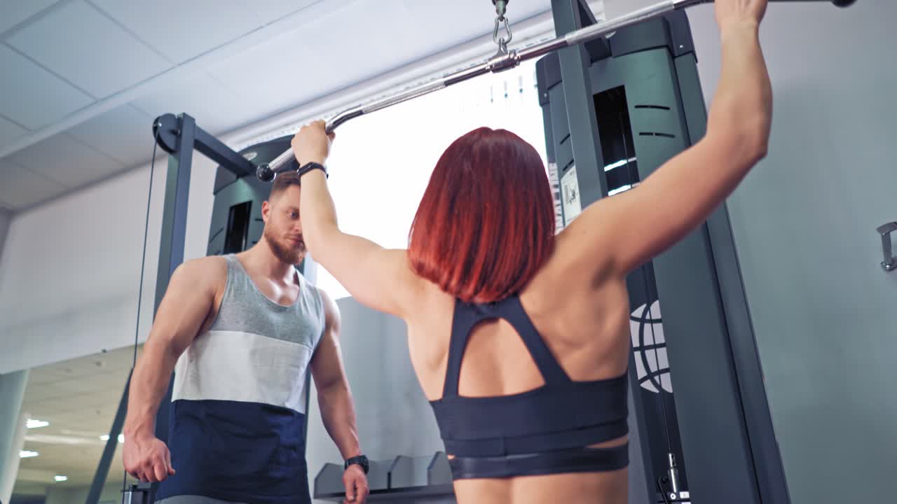 Close-up of female's back working on simulator indoors. Athletic woman is doing exercise while pulling the metal bar down at the fitness centre. Personal trainer man and a sportswoman.