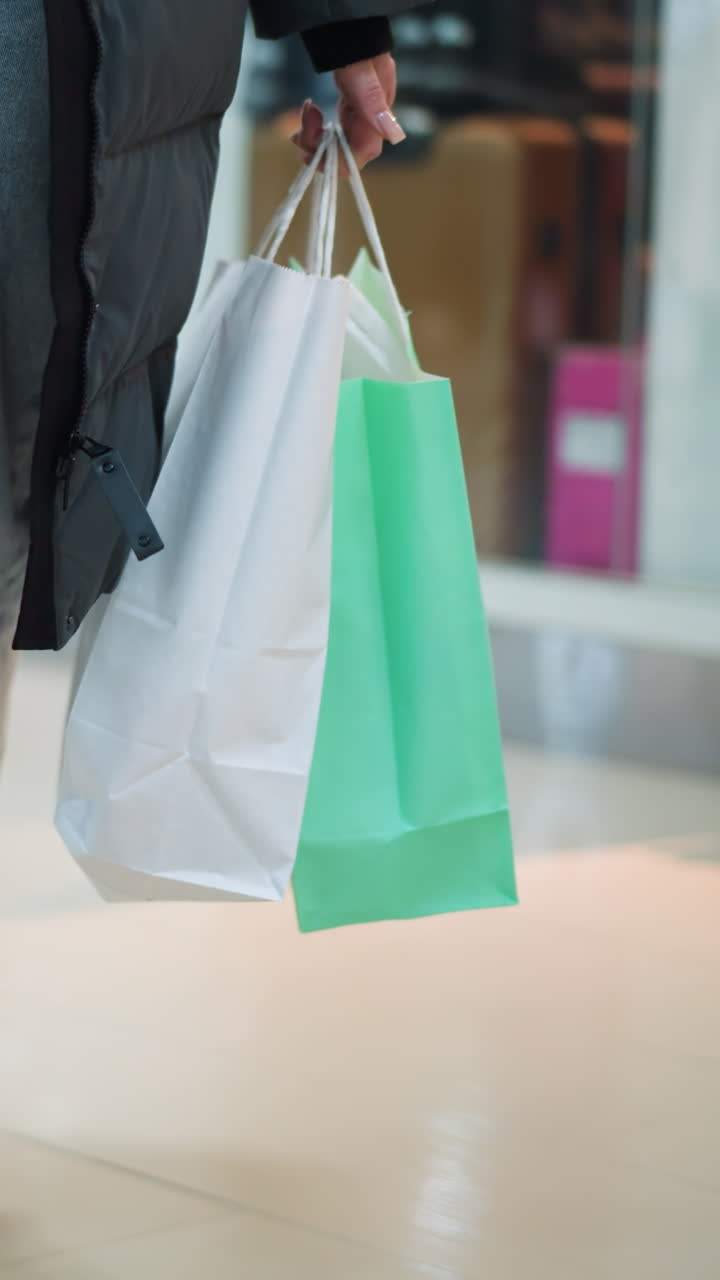 dama en pantalones vaqueros y chaqueta negra caminando en un espacio comercial bien iluminado llevando un bolso negro y dos bolsas de compras, fondo borroso con estantes y exhibiciones de tiendas en un centro comercial moderno