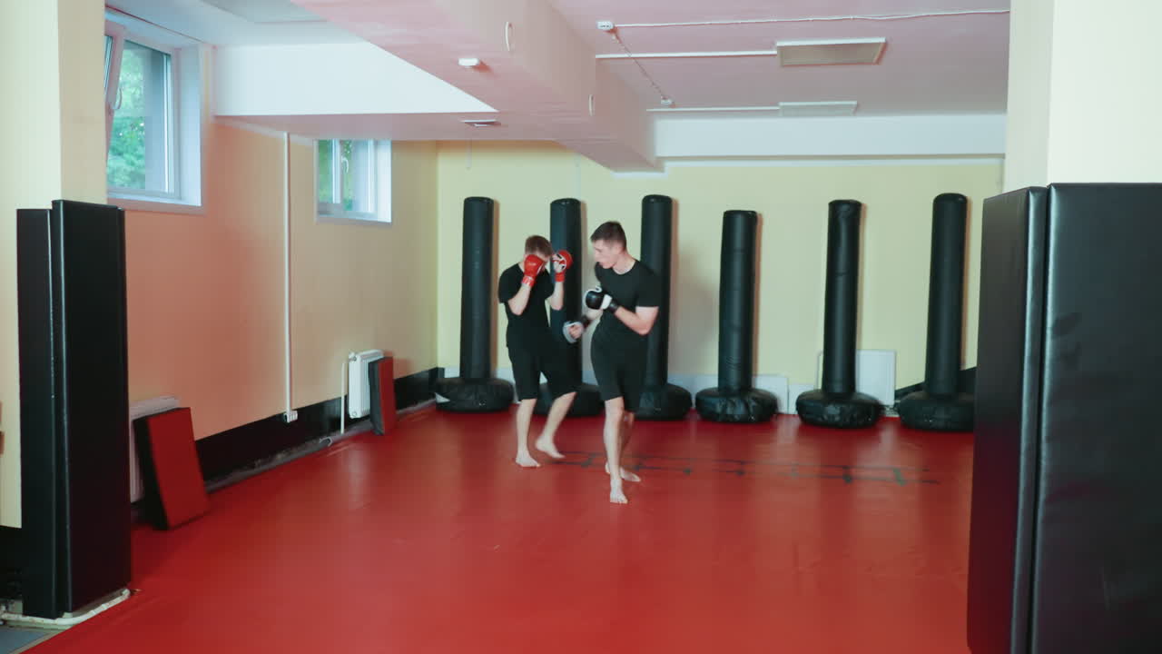 레슬링 선수들이 헬스장 안 빨간색 훈련 매트 위에서 검은색 운동복과 글러브를 착용하고 맨발로 마주 보고 있어요. 노란색으로 칠해진 벽을 배경으로 검은색 펀칭백에 둘러싸여 스파링 세션을 준비하고 있어요.