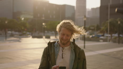 portrait of handsome bearded hipster man texting browsing using smartphone mobile app smiling in windy urban city at sunset