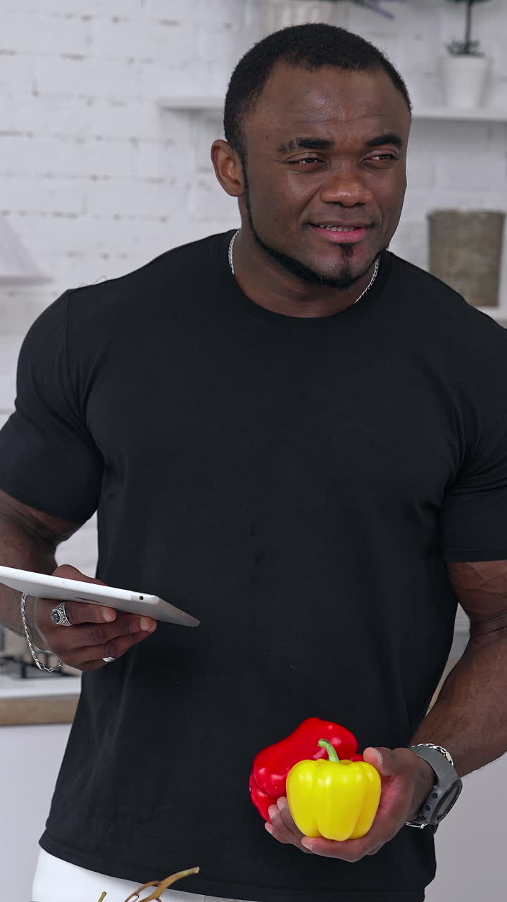 Afro-american sportsman in the kitchen. Portrait of a thoughtful black skinned man with muscular body holding tablet and fresh organic vegetables. Vertical video
