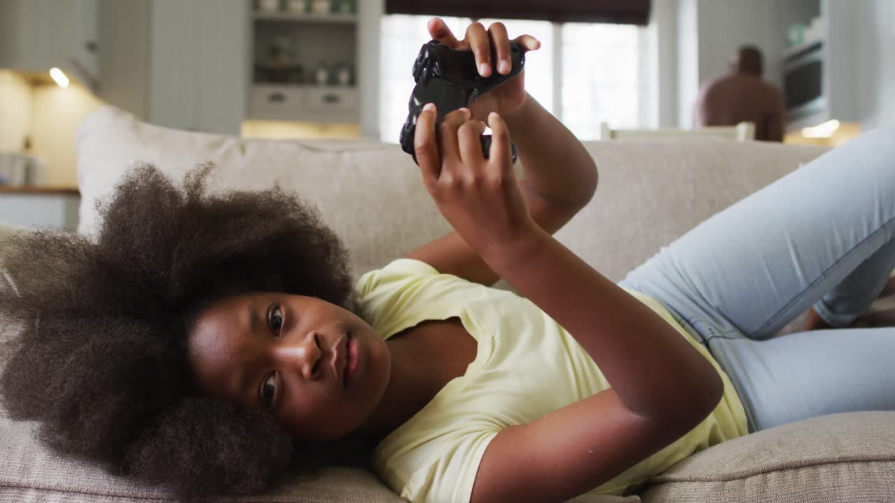 Figlia afroamericana sdraiata sul divano che gioca ai videogiochi con il padre sullo sfondo.