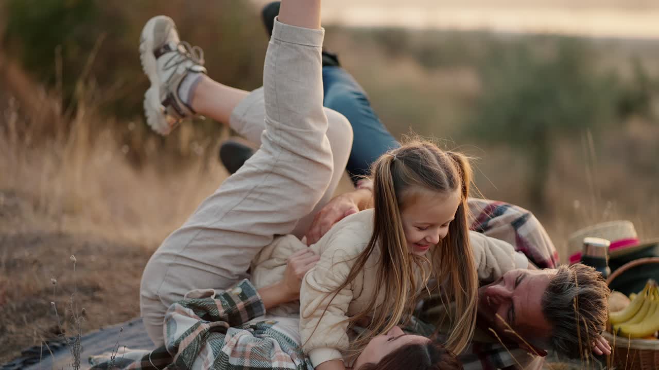 A happy little blonde girl in a white jacket runs to her parents, jumps on them and they fall on the mat during their outdoor recreation and picnic outside the city in the summer. Happy girl runs to hug her parents during a family picnic