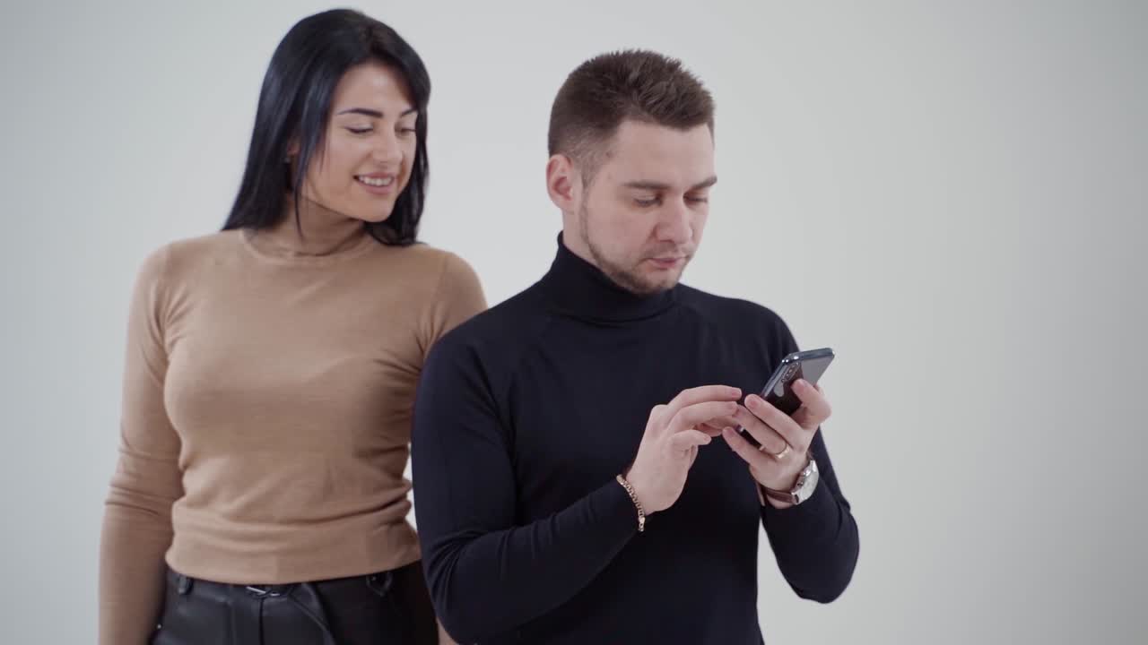 Young happy couple looking at a phone in studio. Man in black sweater holds a cell phone and a woman standing behind him smiling. Slow motion.