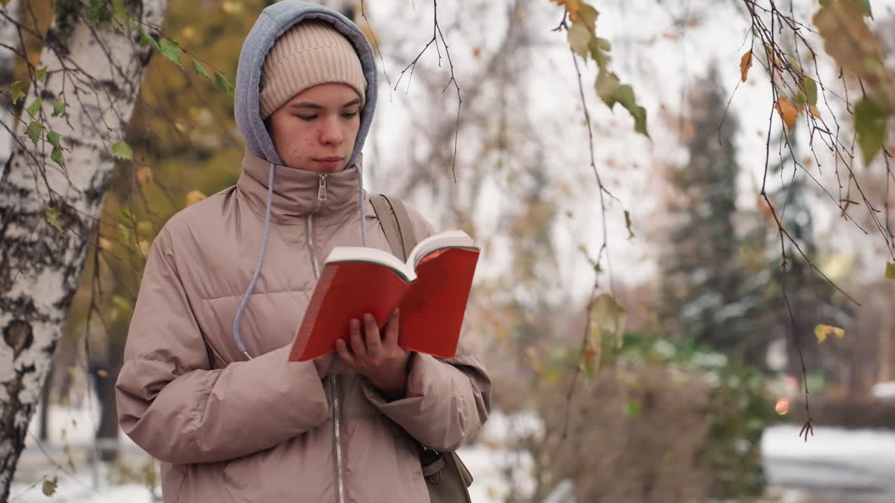 Light skin lady in winter coat and beanie reading red book near birch tree while flipping page in serene autumn weather, with softly blurred background of snow-covered ground and fading foliage