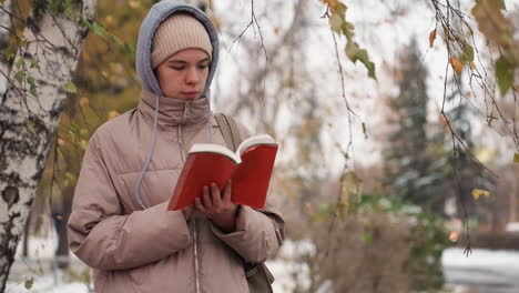 light skin lady in winter coat and beanie reading red book near birch tree while flipping page in serene autumn weather, with softly blurred background of snow-covered ground and fading foliage