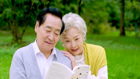 retired asian elderly couple smiling in love embracing grandparents reading book pension leisure on picnic mat in the park