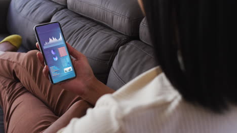 mujer biracial sentada en el sofá usando un teléfono inteligente con estadísticas en la pantalla