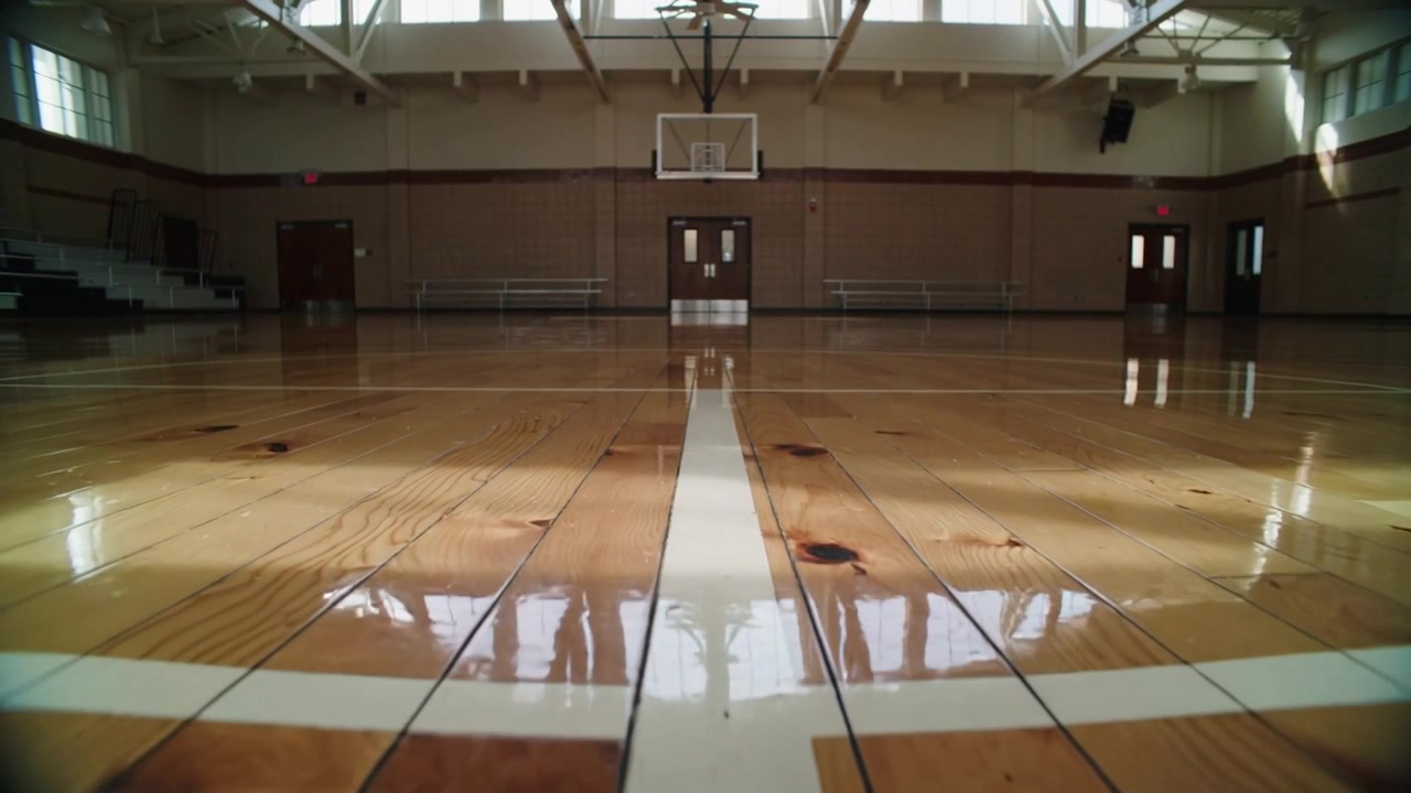 Pulling back camera revealing white center line on hardwood gym floor, showing hoop and windows