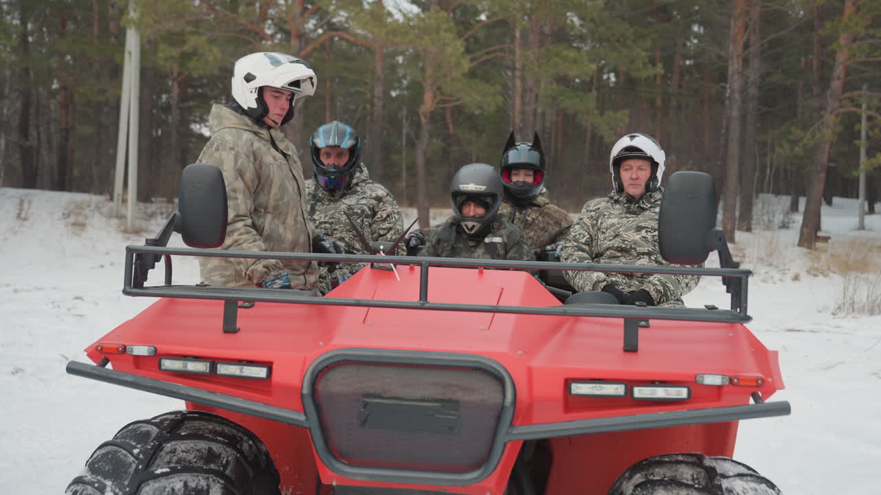 Quattro piloti seduti su un quad rosso in una radura innevata nel bosco. Amici con casco e giacche mimetiche che ridono, pronti per un'avventura fuoristrada, grandi pneumatici e sospensioni robuste visibili, allegro cameratismo.