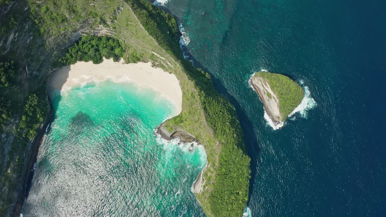 Aerial drone shot in the iconic Kelingking Beach, the 'T-Rex' shaped headland, and the towering limestone cliffs of Nusa Penida. Turquoise Indian Ocean crashes against the rocks and white-sand beach