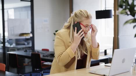 Happy-plus-size-caucasian-casual-businesswoman-using-laptop-and-talking-on-smartphone-at-desk