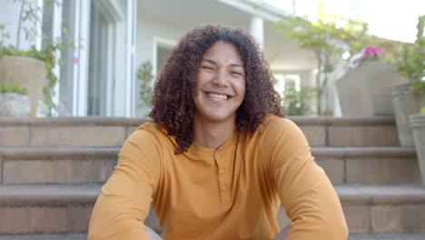 portrait of happy biracial man with long hair sitting on steps smiling in sunny garden, slow motion