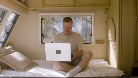 zoom shot of a smiling man sitting on the campervan bed and working on laptop computer