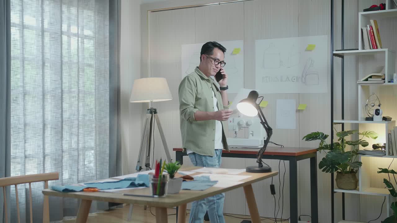 아시아 남자 디자이너가 손에 레이아웃 본드를 보고 사무실에서 고 있는 동안 스마트폰으로 이야기하고 있습니다.