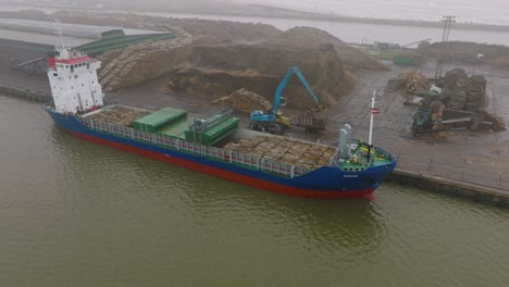 aerial establishing view of wood terminal crane loading timber into the cargo ship, port of liepaja , lumber log export, overcast day with fog and mist, wide distant orbiting drone shot