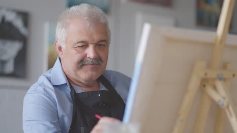 an elderly man in an apron paints a picture with a brush while sitting at the table