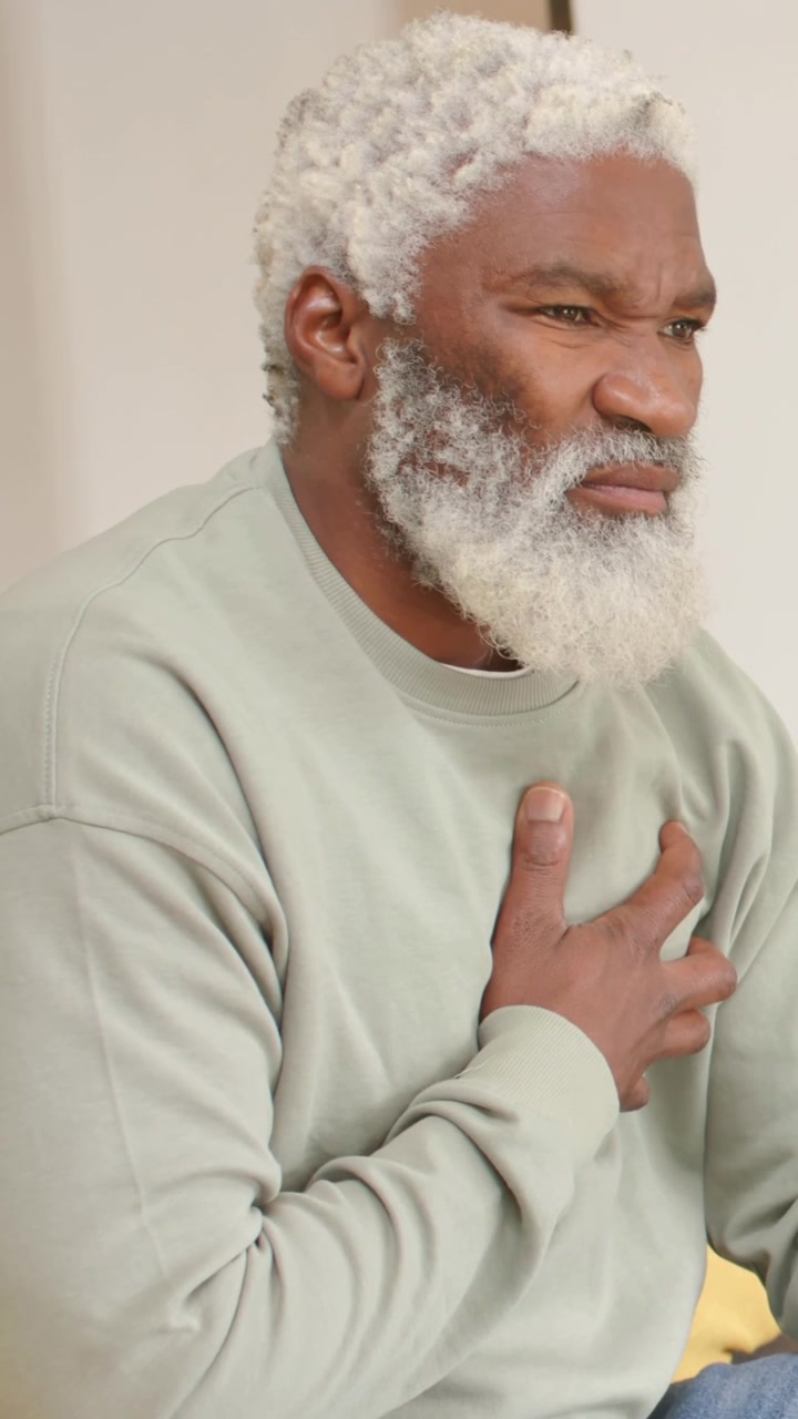 Vertical video: Feeling pain, man in sage top holding left hand on chest on couch, yellow cushion