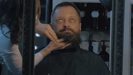 close up view of female barber taking cares of lush beard of man barber applying cosmetics