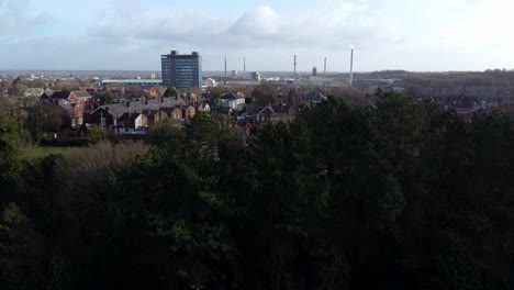 Aerial-view-over-park-trees-to-industrial-townscape-with-blue-skyscraper-on-skyline,-Merseyside,-England