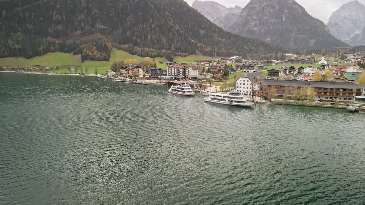 A scenic drone shot at Achensee Pertisau, moving left and ascending. The shot captures passenger boats below, turquoise waters, and majestic mountains in the background.