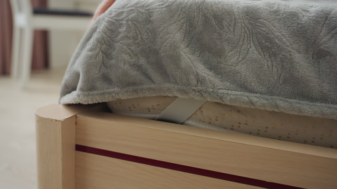 CloseUp Bed Corner With Blanket Showing Mattress Strap And Wooden Frame, Soft Gray Fleece Throw, Gentle Natural Light, Tidy Minimal Bedroom Scene, Focus On Textures And Home Comfort