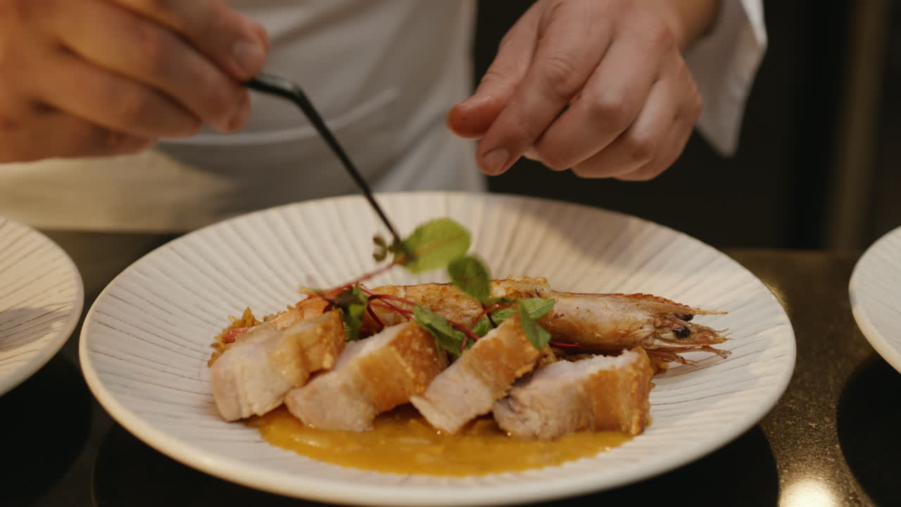 A chef carefully plates crispy pork belly slices over a sauce and prawn, garnishing with fresh herbs using tongs