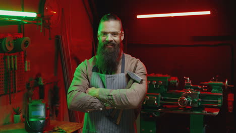 retrato de un soldador caucásico con barba larga sosteniendo un martillo y sonriendo a la cámara en el taller