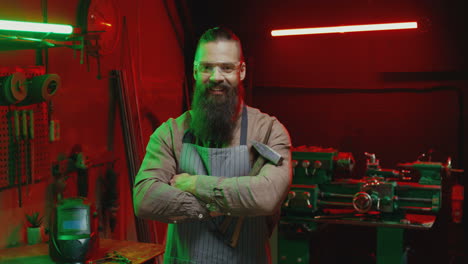 portrait shot of caucasian welder man with long beard holding a hammer and smiling at camera in workshop