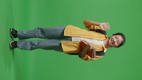 full body of asian woman student with a backpack and some books smiling and waving hand saying bye while standing in the green screen background studio