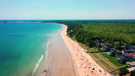 scenic drone video shows saco, maine beaches during peak summer. crowds relax on warm sand, ocean waves crash nearby, and vacation houses dot the coast. a beautiful east coast paradise full of life.