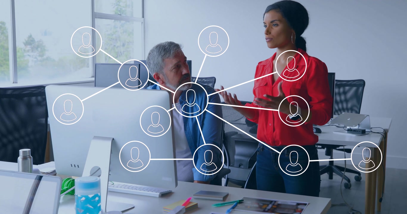 standing woman gesturing at desktop monitor in open-plan office, showing animated network icons
