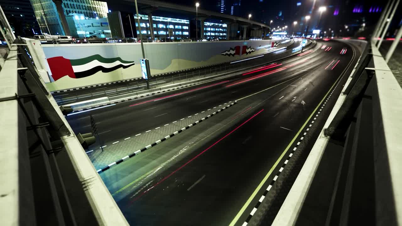 Time-lapse optagelse af en flersporet motorvej i De Forenede Arabiske Emirater om natten, med dynamiske lysspor fra kørende køretøjer. Væggene langs vejen er dekoreret med et vægmaleri af UAE.