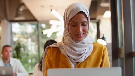 Mature-Businesswoman-Wearing-Headscarf-Working-At-Desk-In-Office-Pausing-To-Look-Out-Of-Window