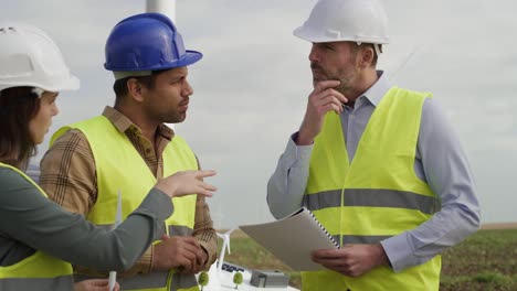 Team-of-caucasian-and-latin-engineers-standing-on-wind-turbine-field-and-discussing-over-plastic-models.