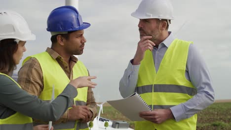 equipo de ingenieros caucásicos y latinos de pie en el campo de turbinas eólicas y discutiendo sobre modelos de plástico.