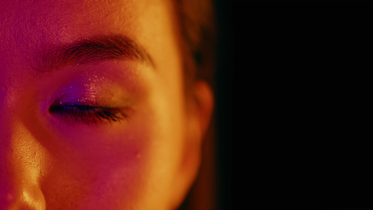 Close-up portrait of a woman's eye and face, lit with vibrant orange and pink neon light.
