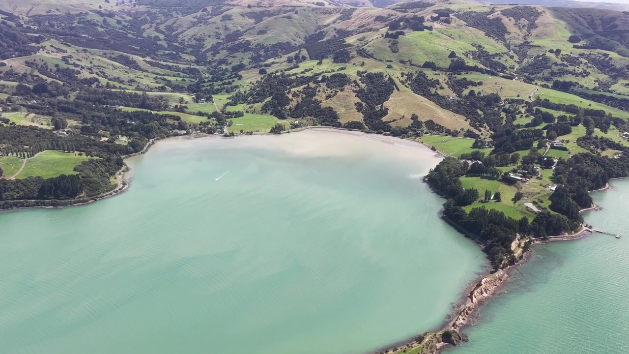 Luftaufnahme einer türkisfarbenen Bucht und sanften grünen Hügeln auf der Südinsel Neuseelands, die eine zerklüftete Küste, klares Wasser und malerische Landschaften zeigt.