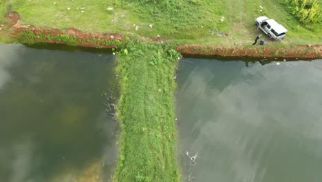 Luftaufnahmen-Vom-Wasser-Zum-Land,-Die-Einen-Landanleger,-Einen-Lastwagen-Und-Zwei-Menschen-Beim-Angeln,-Gras-Und-Unbefestigte-Straße,-Muak-Klek,-Saraburi,-Thailand,-Zeigen