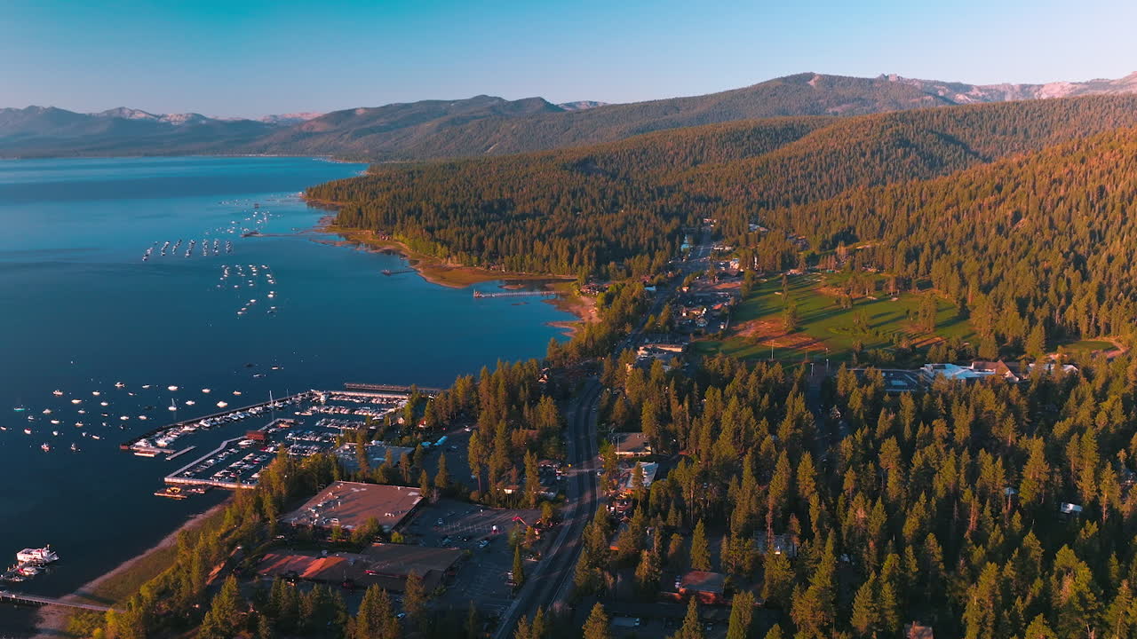Inhabited shore of Lake Tahoe surrounded by thick pine tree forest. Boat pier with lots of yachts from aerial perspective.