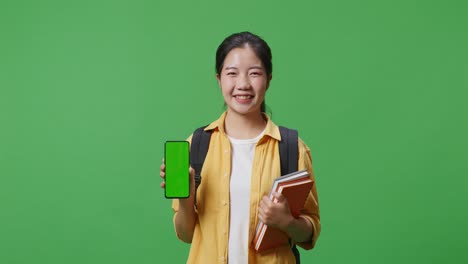 asian woman student with a backpack and some books smiling and showing green screen smartphone to camera while standing in the green screen background studio