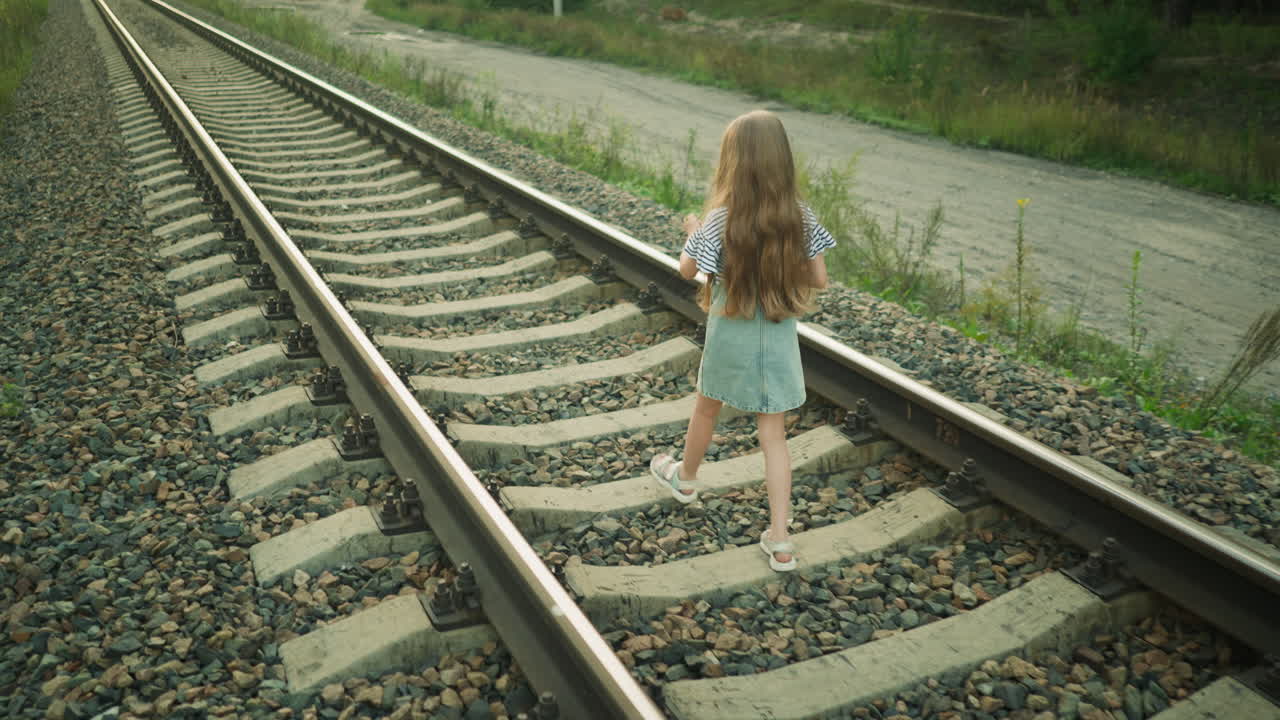 Back view of young girl walking on rail track in rural setting holding object in hand, dressed in denim outfit with long hair flowing, surrounded by gravel stones, grass, and countryside road