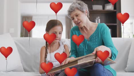 animation of heart balloons over happy caucasian grandmother and granddaughter reading at home