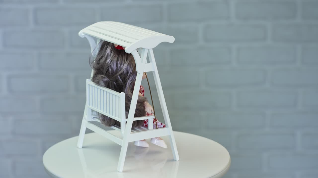 Tiny beautiful doll sitting on the white wooden swing. Beautiful miniature at the rotating stand. Close up.