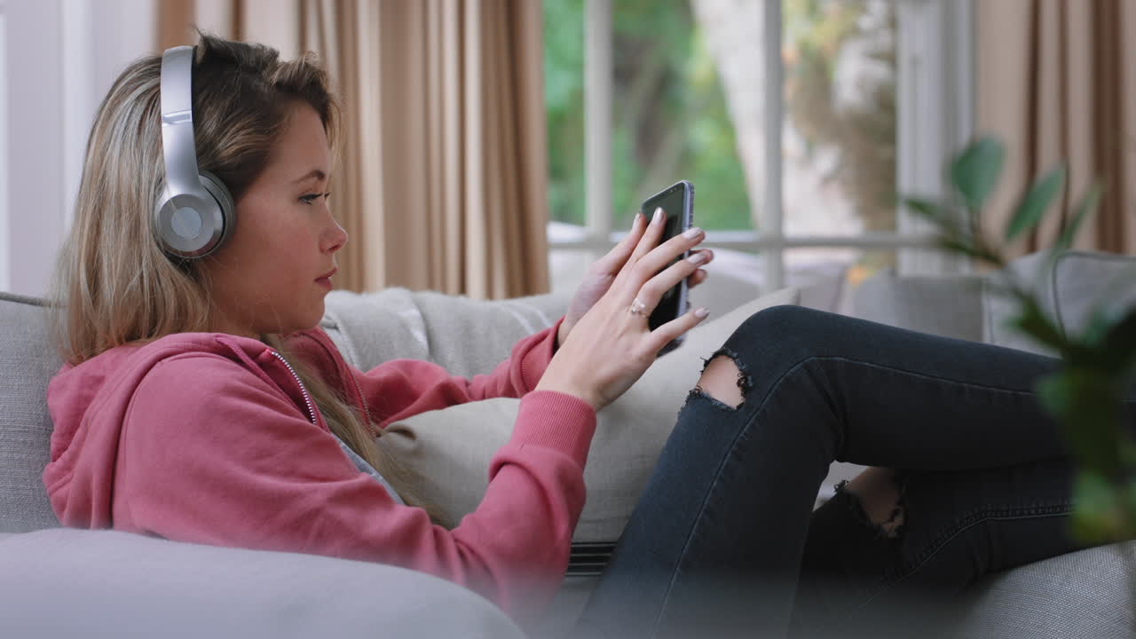 happy teenage girl listening to music wearing headphones relaxing on sofa at home texting with smartphone enjoying sharing lifestyle on social media
