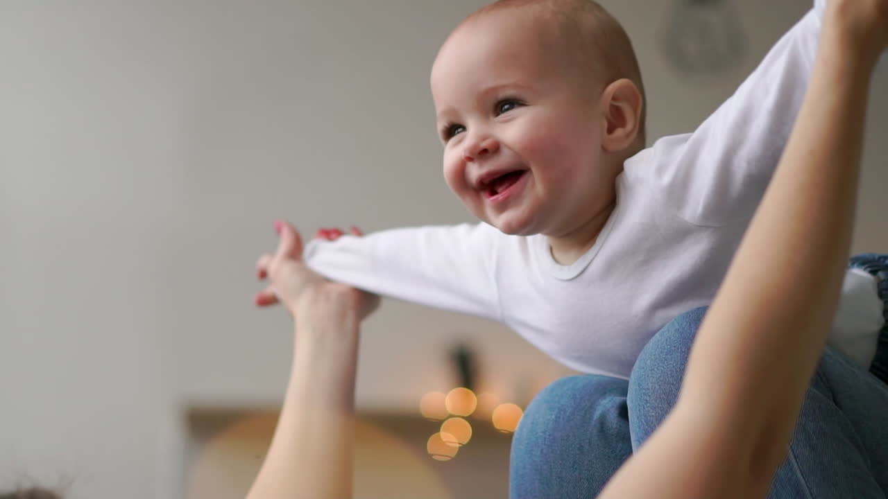 A baby in a white t-shirt lies on his mother's lap imitating the flight of an airplane
