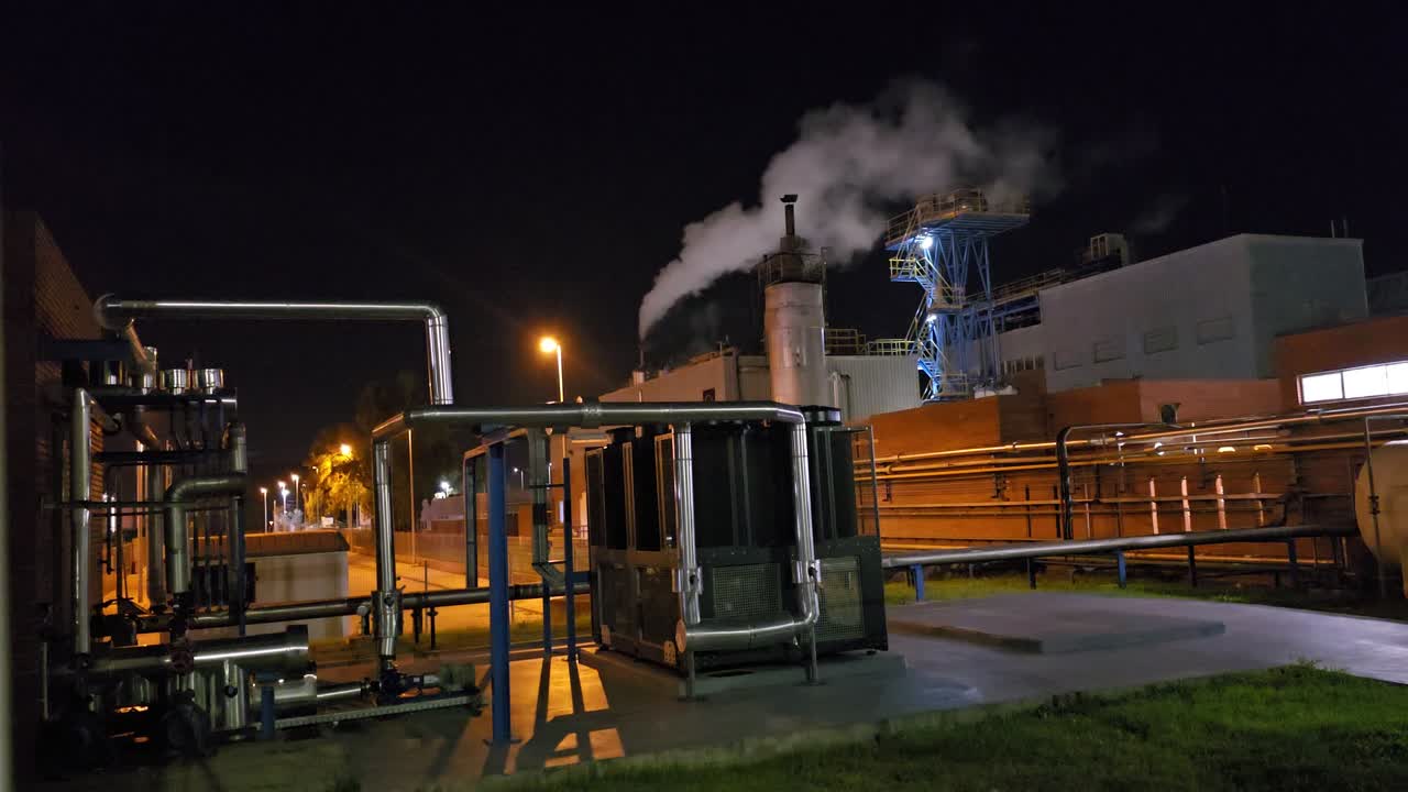 General shot at night in a chemical factory with time laps of starry sky and steam chimney releasing steam