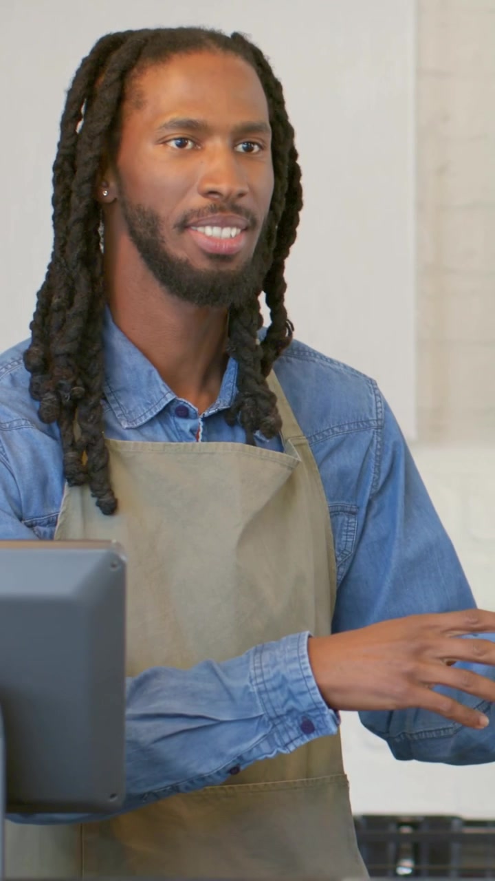 Vídeo vertical: Barista de avental em um café respondendo ao cliente, tocando no PDV e entregando um copo de papel.